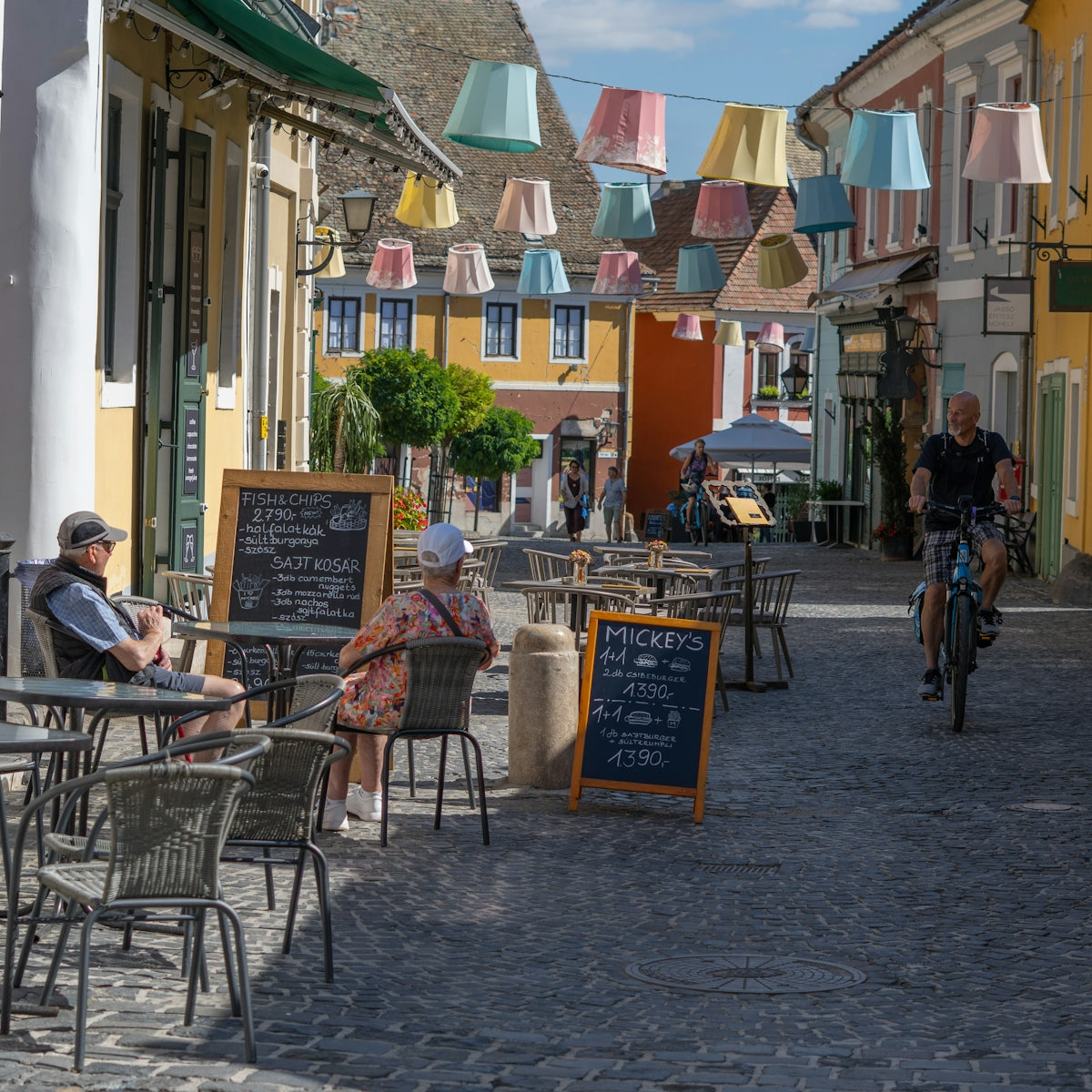 People sit at outdoor cafe tables on cobblestone street